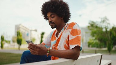 Parktaki bankta akıllı telefonuyla oturan, gömlek giyen Afro-Amerikan bir adamın yakın plan portresi. Yaşam tarzı kavramı. 