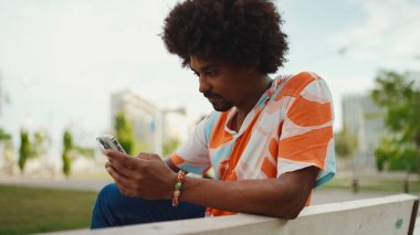 Parktaki bankta akıllı telefonuyla oturan, gömlek giyen Afro-Amerikan bir adamın yakın plan portresi. Yaşam tarzı kavramı. 