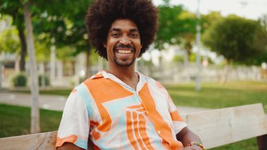 Parktaki bankta rahat bir şekilde kameraya bakarak oturan gülümseyen Afro-Amerikan bir adamın yakın plan portresi. Yaşam tarzı konsepti.