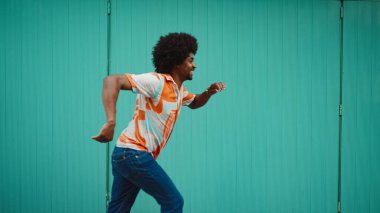 Tişört giyen, kablosuz kulaklıkla müzik dinleyen ve açık mavi duvar arkasında dans eden neşeli, genç Afrikalı Amerikalı adamın yakın çekimi. Yaşam tarzı kavramı. 