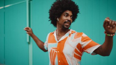 Tişört giyen, kablosuz kulaklıkla müzik dinleyen ve açık mavi duvar arkasında dans eden neşeli, genç Afrikalı Amerikalı adamın yakın çekimi. Yaşam tarzı kavramı. 