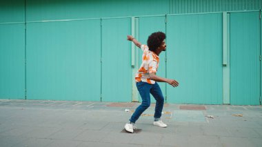 Tişört giyen, kablosuz kulaklıkla müzik dinleyen ve açık mavi duvar arkasında dans eden neşeli, genç Afrikalı Amerikalı adamın yakın çekimi. Yaşam tarzı kavramı. 