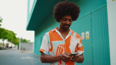 Sosyal ağlarda cep telefonuyla sohbet eden, açık mavi duvarlı arka planda yürüyen neşeli, Afro-Amerikalı genç bir adam. Yaşam tarzı kavramı.