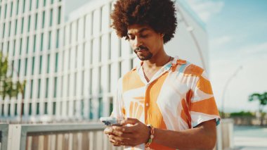 Yakından bakınca, tişört giyen Afro-Amerikalı genç adam sosyal ağlarda şehir arka planında cep telefonuyla yazıyor. Kamera kişiye doğru ilerliyor.. 