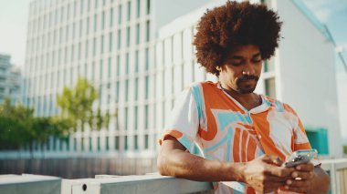 Yakından bakınca, tişört giyen Afro-Amerikalı genç adam sosyal ağlarda şehir arka planında cep telefonuyla yazıyor. Kamera kişiye doğru ilerliyor.. 