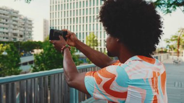 Genç Afro-Amerikan tişört çekimine yakın çekim. Şehir arka planındaki köprüde akıllı telefon tribünlerini kullanın. Yaşam tarzı kavramı