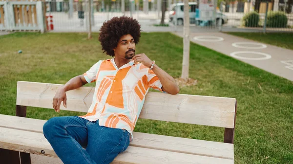 Parktaki bankta rahatça oturan, gömlek giyen Afro-Amerikalı genç adam başka tarafa bakıyor. Yaşam tarzı konsepti. 