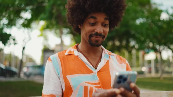Close-up of young African American man wearing shirt sitting on park bench using his smartphone. Closeup of smiling african american man looking at photo video on mobile phone. Lifestyle concept