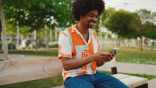 Parktaki bankta oturan Afro-Amerikan gömlekli genç adam akıllı telefonunu kullanıyor. Gülümseyen Afro-Amerikan bir adamın cep telefonundaki fotoğraf videosuna yakından bakması. Yaşam tarzı kavramı
