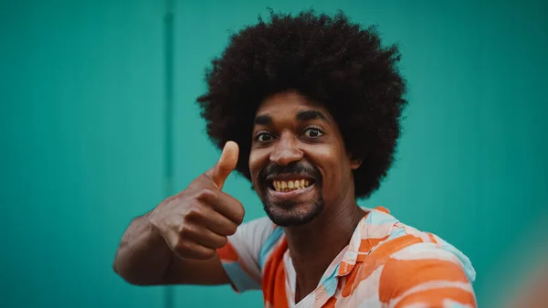 Close-up frontal portrait of cheerful young African American man wearing shirt smiling at the camera against light blue wall background. Lifestyle concept.