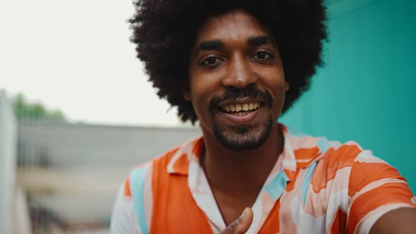 Close-up frontal portrait of cheerful young African American man wearing shirt smiling at the camera against light blue wall background. Lifestyle concept.