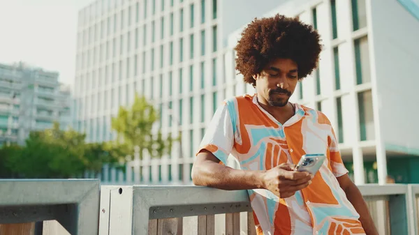 Yakından bakınca, tişört giyen Afro-Amerikalı genç adam sosyal ağlarda şehir arka planında cep telefonuyla yazıyor. Kamera kişiye doğru ilerliyor.. 