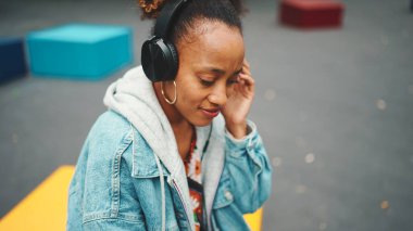 Yakın plan, atkuyruklu, kot ceketli, ulusal desenli, kulaklıklı, modern bina arka planında müzik dinleyen sevimli Afrikalı kız.. 