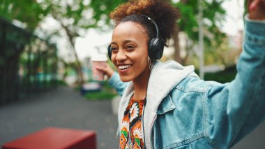 Kot ceketli, gülen Afrikalı bir kız. Üstünde ulusal modelle sokakta yürüyor. Kulaklıkla müzik dinliyor ve dans ediyor.