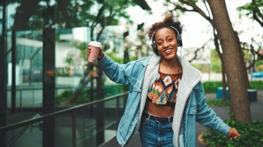 Kot ceketli, gülen Afrikalı bir kız. Üstünde ulusal modelle sokakta yürüyor. Kulaklıkla müzik dinliyor ve dans ediyor.