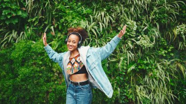 Yakından, kot ceket giyen gülümseyen Afrikalı kız, ekin giyip, milli şablon yürüyüşleriyle şehir parkında yürüyor kulaklıkla müzik dinliyor ve yeşil ağaçlarda dans ediyor.. 