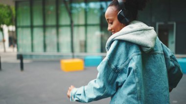 Kot ceketli, gülen Afrikalı bir kız. Üstünde ulusal modelle sokakta yürüyor. Kulaklıkla müzik dinliyor ve dans ediyor.