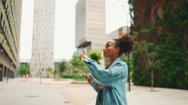 Kot ceket giyen yakın plan Afrikalı bir kız, ulusal desenli ekinlerle modern şehrin sokaklarında dolaşır ve elinde akıllı telefonla şehrin elektronik haritası olan telefon ekranına bakar.