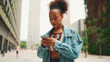 Kot ceket giyen yakın plan Afrikalı bir kız, ulusal desenli ekinlerle modern şehrin sokaklarında dolaşır ve elinde akıllı telefonla şehrin elektronik haritası olan telefon ekranına bakar.