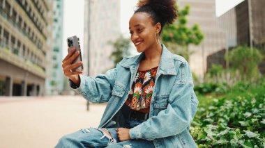Yakın plan portresi at kuyruklu, kot ceket giyen sevimli Afrikalı kız akış yapar, modern şehir arka planında akıllı telefondan video çağrısı yapar.. 