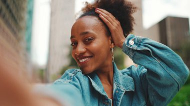 Yakın plan portresi at kuyruklu, kot ceket giyen sevimli Afrikalı kız akış yapar, modern şehir arka planında akıllı telefondan video çağrısı yapar.. 