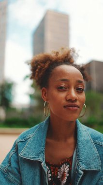 Yakından, at kuyruklu, kot ceketli, ulusal desenli ekili şirin Afrikalı kız otobüs durağında duruyor ve otobüs tarifesine bakıyor..