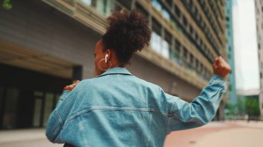 Yakından, at kuyruklu, kot ceketli, milli modelli giyinmiş kulaklıkla müzik dinleyen ve dışarıda dans eden Afrikalı kız.. 