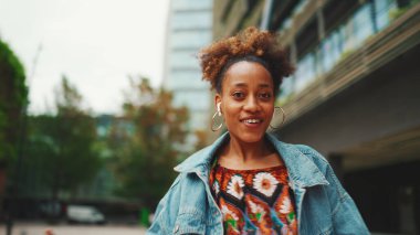 Yakından, at kuyruklu, kot ceketli, ulusal desenli ekili şirin Afrikalı kız otobüs durağında duruyor ve otobüs tarifesine bakıyor..