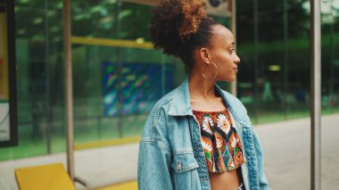 At kuyruklu, kot ceketli, milli desenli ekili şirin Afrikalı kız otobüs durağında duruyor ve gülümsüyor.