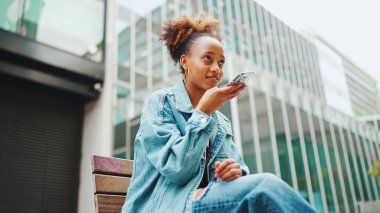 At kuyruklu tatlı Afrikalı kız, kot ceket giyiyor, milli desenli tarlada, bankta oturuyor ve modern binalarda akıllı telefon kullanıyor..
