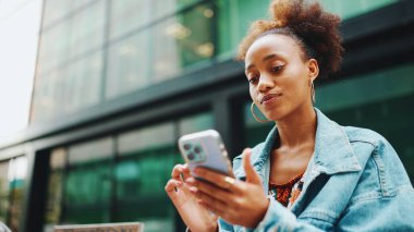 At kuyruklu tatlı Afrikalı kız, kot ceket giyiyor, milli desenli tarlada, bankta oturuyor ve modern binalarda akıllı telefon kullanıyor..