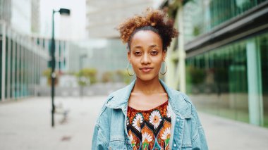 At kuyruklu Afrikalı bir kızın ön portresi. Kot ceket giyiyor. Üzerinde ulusal bir desen ve kameraya bakıp gülümsüyor..