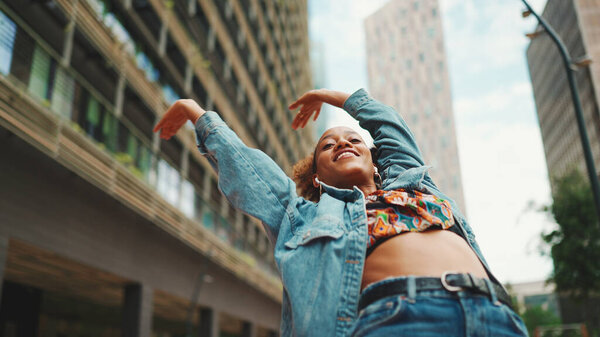 Closeup, smiling African girl with ponytail wearing denim jacket, in crop top with national pattern listening to music on headphones and dancing outdoors. 