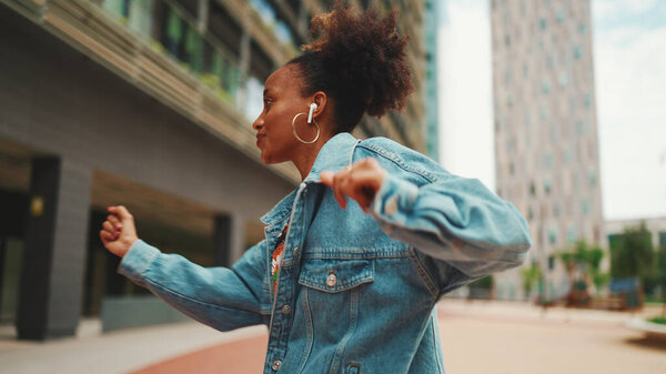 Closeup, smiling African girl with ponytail wearing denim jacket, in crop top with national pattern listening to music on headphones and dancing outdoors. 
