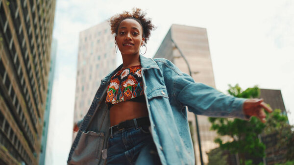 Closeup, smiling African girl with ponytail wearing denim jacket, in crop top with national pattern listening to music on headphones and dancing outdoors. 