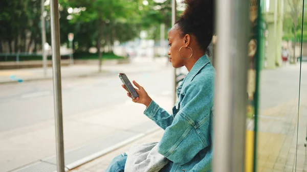 At kuyruklu, kot ceket giyen, milli şablonu olan şirin Afrikalı kız otobüs durağında oturuyor ve cep telefonunu kullanıyor.. 