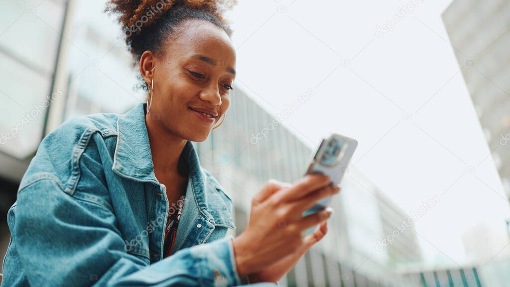 Cute African girl with ponytail, wearing denim jacket, in crop top with national pattern, walking down the street with phone in her hand against modern buildings background. 