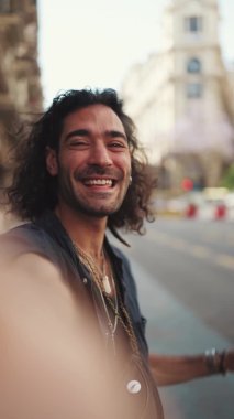 Uzun kıvırcık saçlı ve sakallı genç bir İtalyan sokakta dururken cep telefonundan selfie çekiyor. Akıllı telefonunun kamerasına gülümseyen şık bir adam.