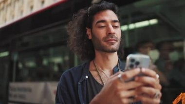 Uzun kıvırcık saçlı ve sakallı çekici İtalyan çocuk cep telefonu kullanıyor. Yaklaşan bir otobüsün arka planında akıllı telefondan mesaj yazan şık bir adam.