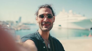 Uzun kıvırcık saçlı ve sakallı genç İtalyan adam cep telefonundan selfie çekiyor. Güneş gözlüklü şık adam yat arka planındaki akıllı telefon setinin kamerasına gülümsüyor.