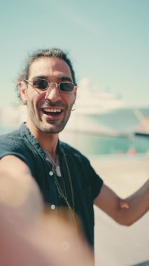 Clouse-up, uzun kıvırcık saçlı ve sakallı genç İtalyan cep telefonundan selfie çekiyor. Güneş gözlüklü şık bir adam telefonuna gülümsüyor ve yatını işaret ediyor.