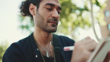 Yakın plan, at kuyruklu ve sakallı genç İtalyan adam sokak bankında oturur ve şehir planındaki kağıt üzerine kalemle çizimler yapar.