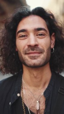 Uzun kıvırcık saçlı ve sakallı çekici İtalyan genç adam kameraya bakıyor ve eski binaların arka planına gülümsüyor. Kulağında küpe ve bir sürü zincir olan şık bir adam.