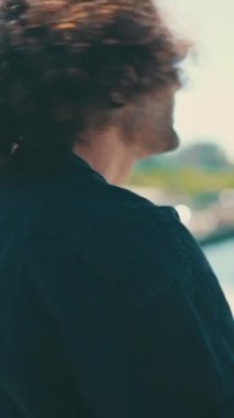 Uzun kıvırcık saçlı ve güneş gözlüklü sakallı genç İtalyan limanda duruyor ve gemilere ve yatlara bakıyor.