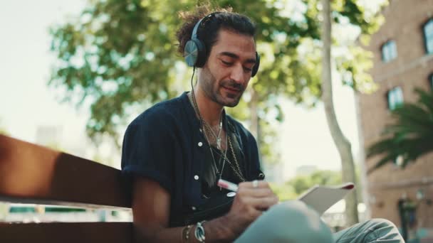Młody Włoch Kucykiem Zarostem Siedzi Słuchawkami Ławce Ulicznej Słucha Muzyki — Wideo stockowe