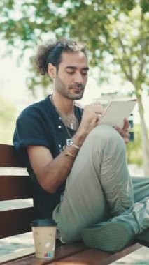 At kuyruklu ve sakallı genç İtalyan adam sokak bankında oturuyor ve kağıt üzerine kalemle çizimler yapıyor.