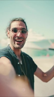 Clouse-up, uzun kıvırcık saçlı ve sakallı genç İtalyan cep telefonundan selfie çekiyor. Güneş gözlüklü şık bir adam telefonuna gülümsüyor ve yatını işaret ediyor.