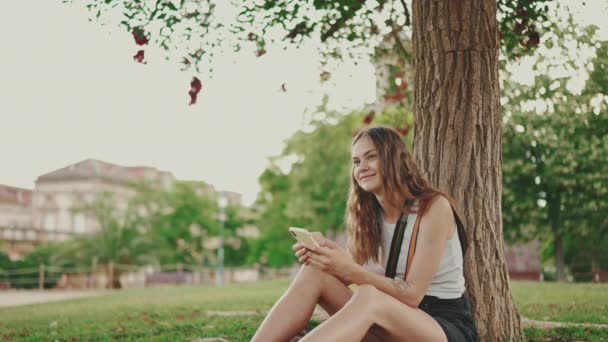 윗옷을 기다란 아름다운 소녀는 의나무 아래푸른 잔디에 전화를 전화기에 사진들을 — 비디오