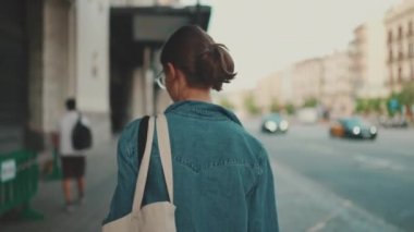 At kuyruklu, kot ceketli, elinde cep telefonu olan genç bir kadın şehir manzarası arka planında sokaklarda yürüyor. Akıllı telefonda gezinen kız bilgileri