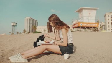 Öğle yemeği vakti. Şirin bir kız kumsalda oturmuş evcil hayvan deneyleri yapıyor. Öğle yemeğini modern şehir arka planında, sahilde yiyor.